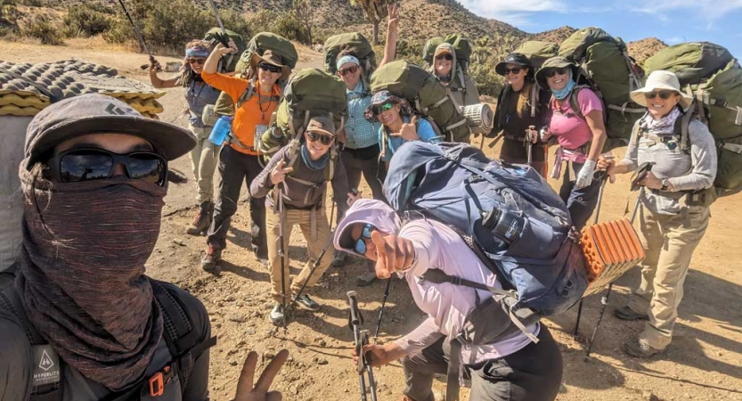 A group of women wearing backpacks stand for a group photo in a desert environment. 